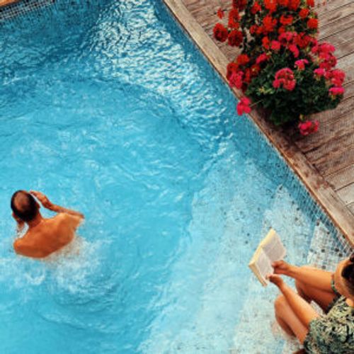 Två personer badar och läser i pool med ljus liner, brun trall och röda pelargoner