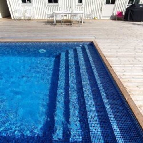 Pool med helgaveltrappa i mörkblå mosaikliner, trädäck och vitt hus i bakgrunden