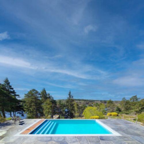 Pool med ljusgrå granitsarg i öppet landskap med tallar och sjöutsikt